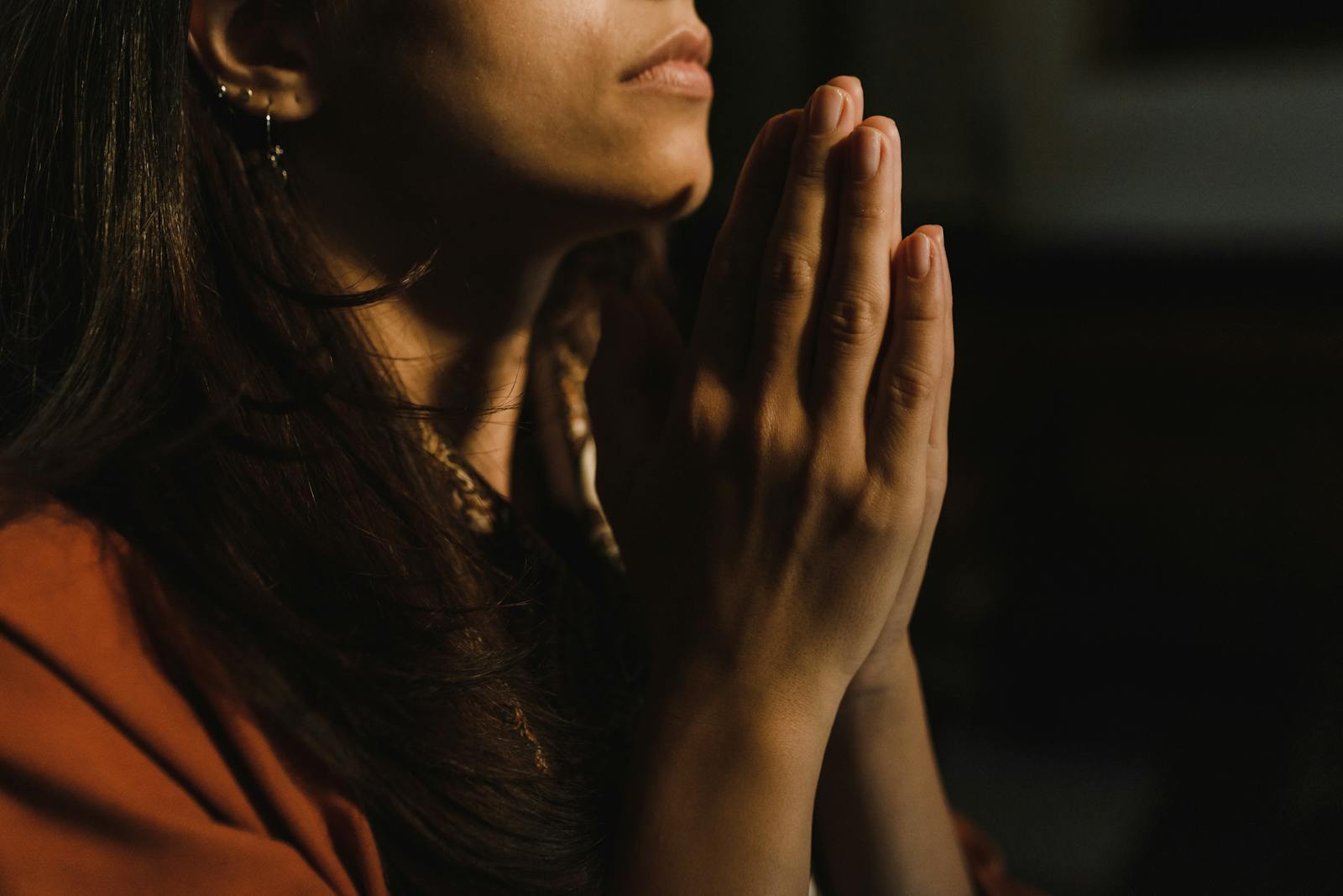 Close-up of a woman with folded hands praying, captured in a serene indoor setting with low lighting.